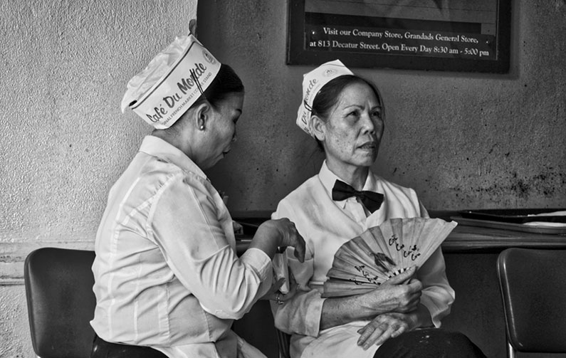 black and white photo of two women in paper hats reading "Cafe Du Monde," one holding fan