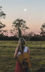 Woman with an acoustic guitar slung over her back facing a moon rise