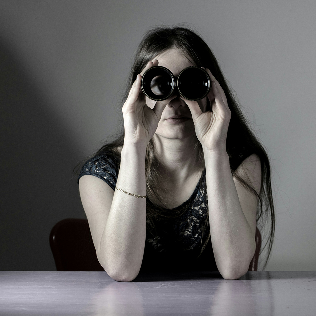 Woman in darkened room looks out at the reader through binoculars.