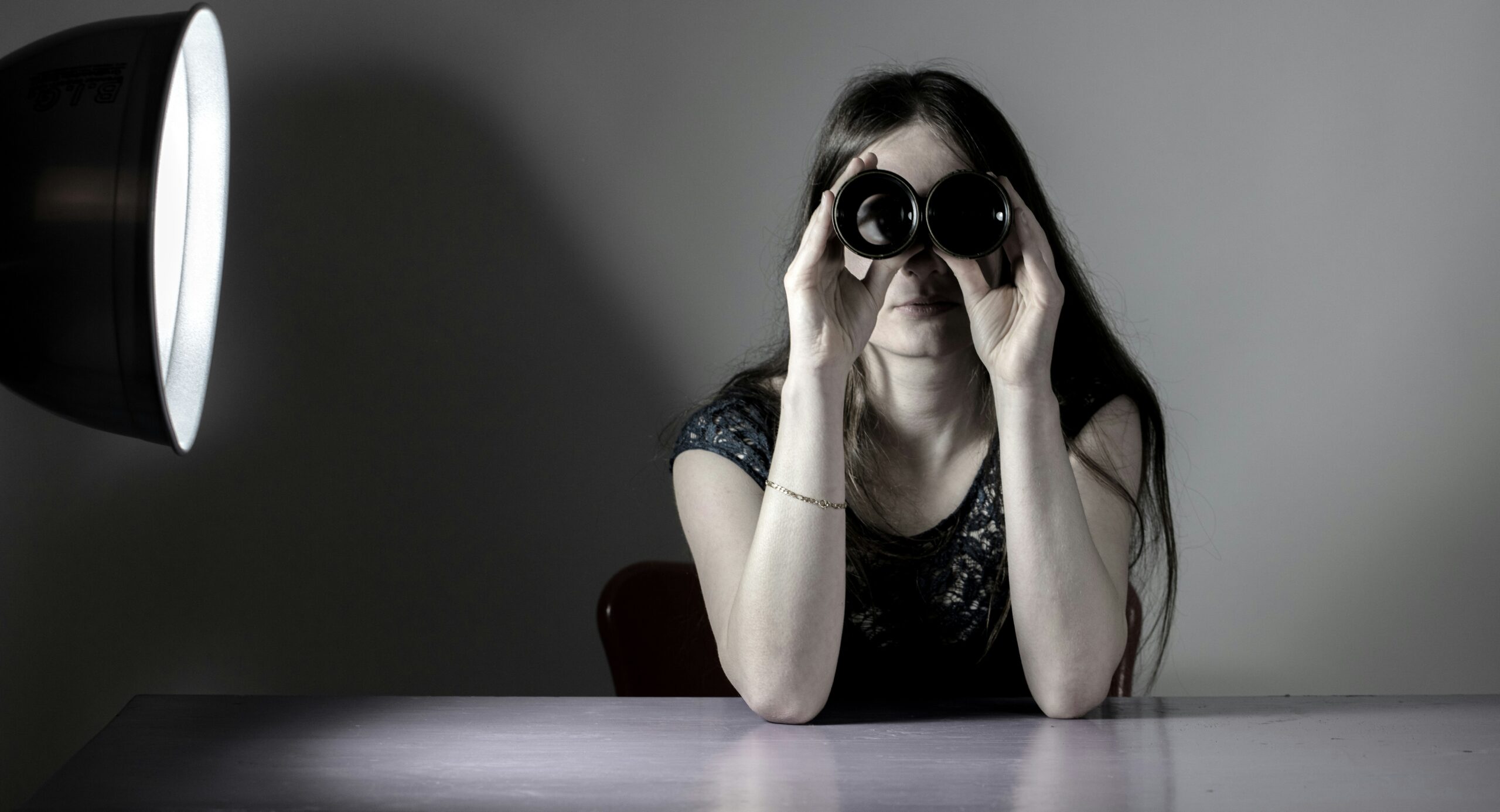 Woman in darkened room looks out at the reader through binoculars.