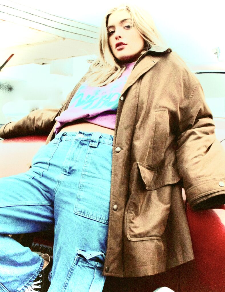 Young Blonde Woman in Cargo Jeans and a Brown Coat Leaning against a Red Car