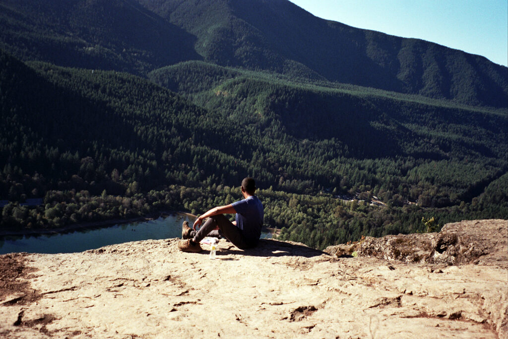 Seated man on mountain top looking out at tree-covered hills and a lake.