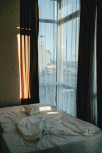 Unmade bed in windowed corner overlooking ocean, palm trees, and another building.