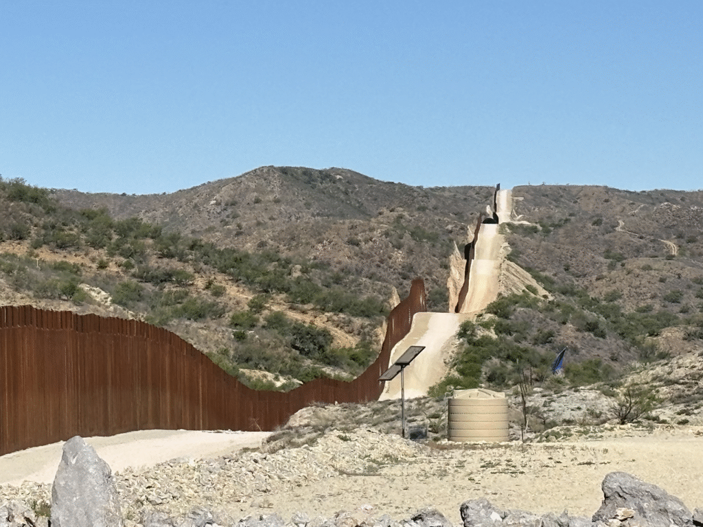 Photo of the border wall stretching away over hills through desert scrub.