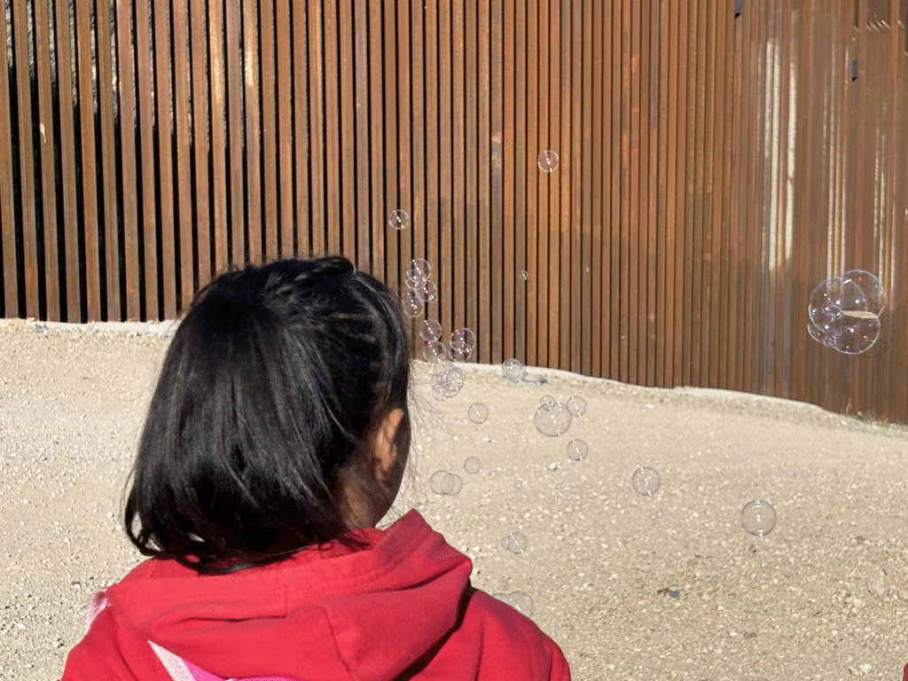 Photo of a young child in a hoodie blowing bubbles with the border wall in the background.